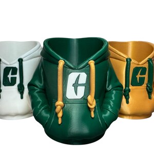 Puede incluir: Tres macetas con forma de sudadera con capucha en blanco, verde y dorado. Cada maceta presenta una capucha con cordones y un logotipo en la parte delantera. La sudadera verde tiene cordones dorados, mientras que la dorada tiene cordones amarillos.
