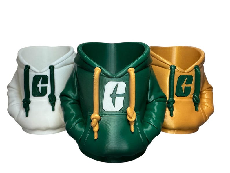 Puede incluir: Tres macetas con forma de sudadera con capucha en blanco, verde y dorado. Cada maceta presenta una capucha con cordones y un logotipo en la parte delantera. La sudadera verde tiene cordones dorados, mientras que la dorada tiene cordones amarillos.