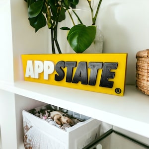 Pode incluir: Uma placa amarela com as palavras "AppState" em letras pretas e brancas. A placa está em uma prateleira branca com uma planta em um vaso transparente atrás.