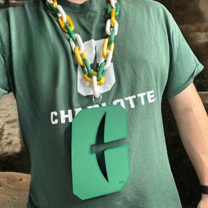 May include: A green and white lanyard with a green plastic charm shaped like the letter "C" with a stylized arrow inside. The lanyard has a white chain with yellow links. The charm has the text "TM" at the bottom.