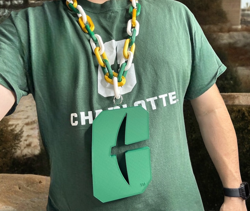 May include: A green and white lanyard with a green plastic charm shaped like the letter "C" with a stylized arrow inside. The lanyard has a white chain with yellow links. The charm has the text "TM" at the bottom.