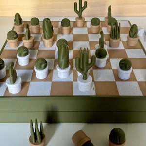 Puede incluir: Un juego de ajedrez verde y marr&oacute;n con piezas con forma de cactus. El tablero est&aacute; cerrado y las piezas est&aacute;n esparcidas a su alrededor.