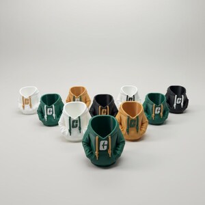 Puede incluir: Una colecci&oacute;n de macetas en miniatura con forma de sudadera con capucha en varios colores, incluyendo blanco, verde, negro y dorado. Cada maceta presenta una capucha y un dise&ntilde;o de bolsillo, con un logotipo en la parte delantera. Las macetas est&aacute;n dispuestas sobre una superficie gris claro.