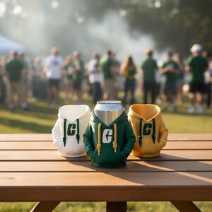 Puede incluir: Tres enfriadores de latas con forma de sudaderas con capucha, en blanco, verde y amarillo, est&aacute;n sobre una mesa de picnic de madera. El enfriador verde contiene una lata plateada. Cada enfriador tiene la letra "G".