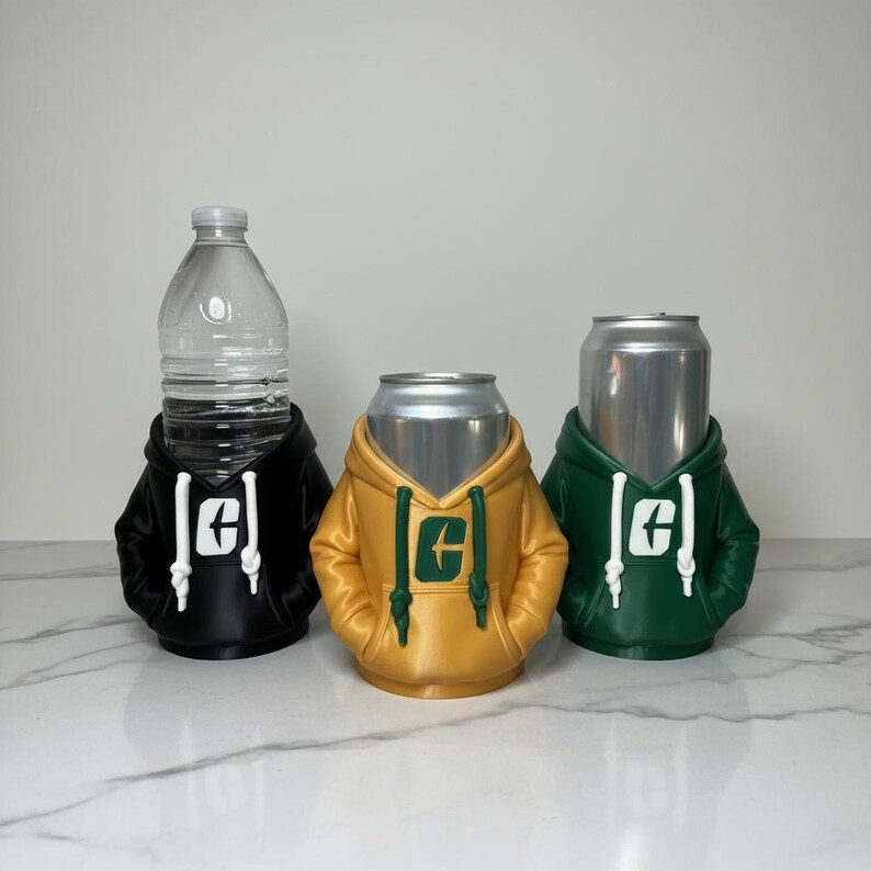 Puede incluir: Tres soportes para bebidas con forma de sudaderas con capucha. Una sudadera negra sostiene una botella de agua, una sudadera amarilla sostiene una lata y una sudadera verde sostiene una lata. Cada sudadera tiene un cord&oacute;n blanco y un logotipo.