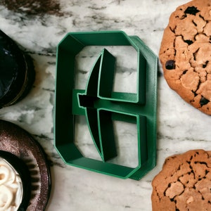 May include: Green plastic cookie cutter shaped like a pickaxe. The cutter is on a marble surface.