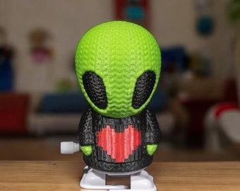 Puede incluir: Un juguete de cuerda verde extraterrestre con un suéter negro y un corazón rojo en la parte delantera. El juguete está sobre una superficie de madera.
