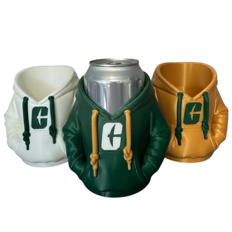Puede incluir: Tres enfriadores de latas con forma de sudaderas con capucha, en blanco, verde y dorado. Cada enfriador tiene una capucha, cordones y un logotipo en la parte delantera. Una lata plateada est&aacute; dentro del enfriador verde. Los enfriadores est&aacute;n dise&ntilde;ados para mantener las bebidas fr&iacute;as.