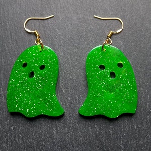 Op de afbeelding: Een paar groene glitter-geestoorbellen met gouden haakjes. De geesten hebben drie gaten voor ogen en een mond.