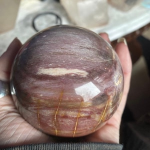 May include: A polished sphere of petrified wood with a swirling pattern of pink, brown, and white. The sphere has thin, gold-colored lines running through it.