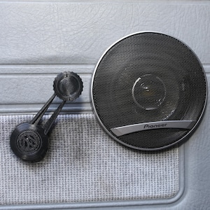 Może przedstawiać: Zbliżenie panelu drzwi samochodu z czarnym głośnikiem Pioneer i czarną korbą do okna. Głośnik jest okrągły z siatkową osłoną i srebrnym logo Pioneer. Korbka do okna jest z czarnego plastiku.