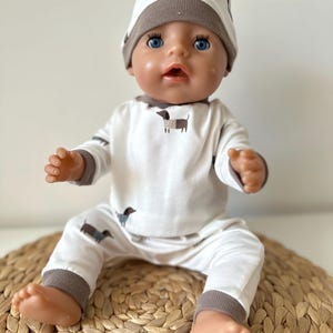 Peut inclure: Une poupée pour bébé vêtue d'un ensemble blanc orné d'un motif de teckel marron. L'ensemble comprend un haut à manches longues, un pantalon et un bonnet. La bordure de l'ensemble et du bonnet est marron. La poupée a les yeux bleus et est assise sur un tapis tissé.