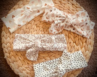May include: An assortment of knotted headbands in various patterns, including hearts, floral, and polka dots. The headbands are in neutral tones of white, cream, and brown, arranged on a woven wicker surface.