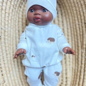 Peut inclure: Une poupée à la peau foncée portant un bonnet blanc, un haut à manches longues et un pantalon assorti. La tenue présente des illustrations de hérissons bruns. La poupée est posée dans un panier en osier.