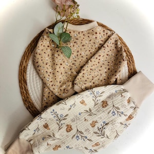 Pode incluir: Um conjunto de bebé bege. A parte de cima tem um padrão floral com pequenas flores castanhas e brancas, e as calças apresentam um estampado de coelhos e flores. A parte de cima tem gola redonda e mangas compridas. As calças têm punhos bege. O conjunto está sobre uma esteira tecida.