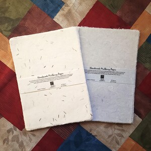 May include: Two handmade mulberry paper notebooks, one white and one light blue, are displayed on a colorful patchwork quilt. Each notebook has a paper band with text. The paper has visible flecks and is made in Thailand.