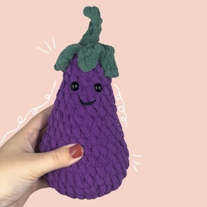 Puede incluir: Un juguete de peluche de berenjena de crochet morado y verde con una cara sonriente. La berenjena está siendo sostenida por una mano.