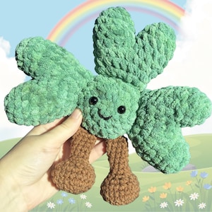 May include: A handmade green crochet clover plush toy with a smiling face and brown legs. The clover has four leaves and is held in a hand. The background features a rainbow and a cartoon landscape.