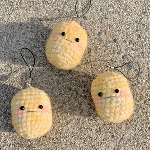 May include: Three handmade, crocheted potato charms with black bead eyes and a small, stitched mouth. Each charm has a loop for hanging and rosy cheeks. The charms are light yellow and sit on a textured, gray surface.
