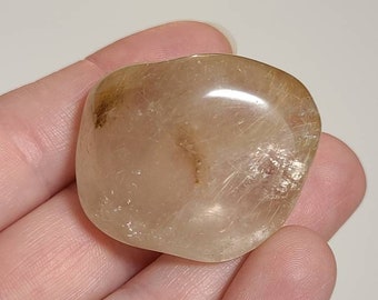 May include: A translucent, light brown, oval-shaped, tumbled stone with a letter A on a white piece of paper.