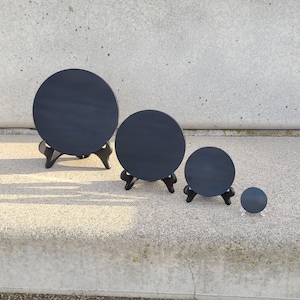 May include: Three black slate round coasters of different sizes on small black stands. The coasters are on a gray concrete surface.