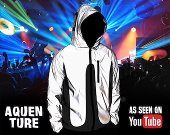 Peut inclure: Une veste à capuche argentée réfléchissante avec des panneaux latéraux noirs et une fermeture éclair noire. Le texte AQUEN TURE est à gauche et le texte AS SEEN ON YouTube est à droite. La veste est présentée sur un fond flou de lumières de scène.