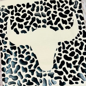 May include: A white fabric with a black cow print design. A white silhouette of a bull's head is centered on the fabric.