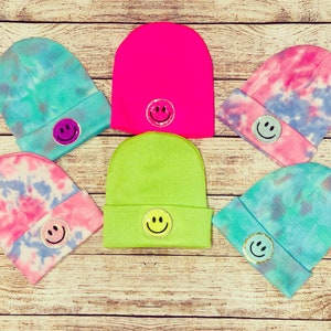 May include: Six colorful knit beanies with tie-dye patterns and smiley face patches. The beanies are in shades of pink, blue, green, and yellow.