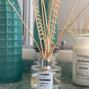 May include: A clear glass diffuser bottle with a silver cap and wooden reeds. The label reads "Mango + Tangerine" and "Made with Love." The bottle is 177 ml and sits on a textured surface. Other teal glass bottles and a candle are in the background.