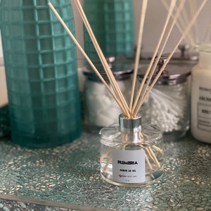 Op de afbeelding: Een heldere glazen aromatherapie-diffuser met houten stokjes. Het label luidt "PLUMERIA" en "PORSH JE CO." met "MADE WITH LOVE" eronder. De diffuser staat op een turquoise en wit gespikkeld oppervlak.