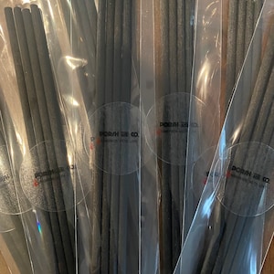 Może przedstawiać: Kilka wiązek ciemnoszarych pałeczek do jedzenia jest zapakowanych w przezroczyste plastikowe rękawy. Każdy pakiet ma okrągłą naklejkę z napisem "PORSH & CO. HANDMADE WITH LOVE". Pałeczki wydają się być wykonane z drewna.