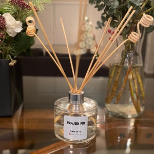 May include: A clear glass diffuser bottle with a silver collar and wooden reeds. The label reads "Fraser Fir" and "Made with Love". The bottle contains a clear liquid and is surrounded by decorative plants.