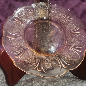 May include: A pink glass saucer with a floral design. The saucer has a scalloped edge and is made of clear glass with a pink tint.