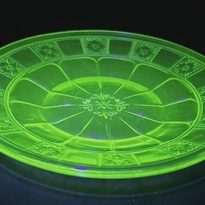 May include: A round, glowing green glass plate with a decorative floral and geometric pattern. The plate has a raised rim and a central design radiating from the center. The plate is approximately 10 inches in diameter.