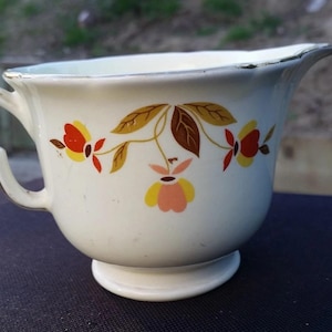 May include: Cream-colored vintage creamer with a floral design in red, yellow, and brown. The creamer has a handle and a spout, and sits on a small pedestal base. The rim and handle are trimmed with gold.