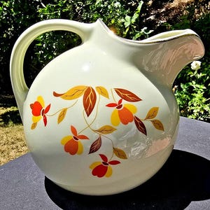 May include: Cream-colored ceramic pitcher with a large handle and a spout. The pitcher is decorated with a floral pattern in shades of orange, yellow, and brown. A thin gold trim accents the rim.