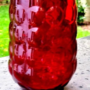 May include: A vibrant red glass tumbler with a textured, bubble-like pattern. The cylindrical shape tapers slightly towards the base. The glass is transparent, allowing light to pass through, and is suitable for beverages.