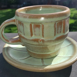 Puede incluir: Un juego de taza y platillo de cerámica verde y marrón con un diseño texturizado y acanalado. La taza tiene un asa y el platillo tiene un borde festoneado.