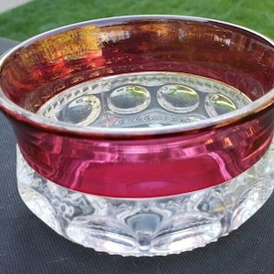 May include: A clear glass bowl with a ruby red rim and a wide band of ruby red around the middle. The base of the bowl is clear with a repeating pattern of oval shapes. The bowl is sitting on a dark surface.