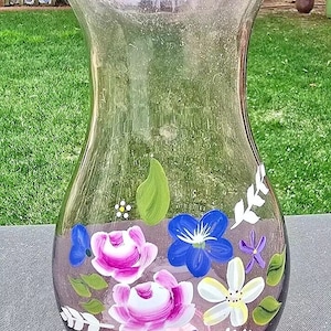 May include: A clear glass vase with a ruffled rim, hand-painted with pink, blue, white, and purple flowers. The vase has a light pink tint.