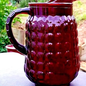 Puede incluir: Una jarra de vidrio rojo rubí con un patrón texturizado similar a burbujas. La jarra tiene un asa y un pico vertedor. Probablemente se utiliza para servir bebidas.