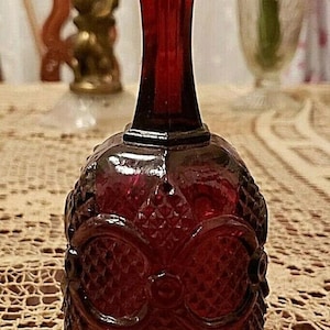 Puede incluir: Una campana de vidrio rojo oscuro con una superficie texturizada y un diseño decorativo. La campana está sentada sobre un mantel de encaje blanco.