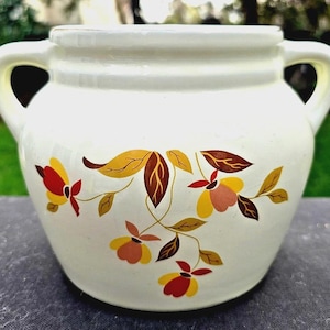 Könnte beinhalten: Cremefarbener Keramiktopf mit zwei Griffen und floralem Design. Der Topf hat eine abgerundete Form mit einem leicht ausgestellten Rand. Das florale Muster umfasst gelbe, rote und braune Blätter und Blüten und verleiht einen Hauch von Vintage-Charme.