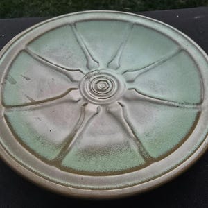 May include: A circular ceramic plate with a light green and brown glaze. The plate has a central spiral design with radiating lines, forming a sunburst pattern. The rim is a darker brown colour.