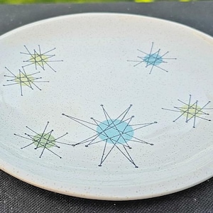 Op de afbeelding: Een ronde, gebroken witte keramische plaat met een gespikkeld oppervlak. De plaat heeft een modern design uit het midden van de eeuw met sterpatronen in blauw, groen en geel, omlijnd in zwart. Een decoratief stuk voor de keuken of eetkamer.
