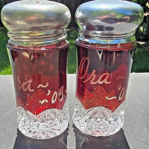 Puede incluir: Dos saleros y pimenteros vintage de vidrio con tapas plateadas. Los saleros están llenos de un líquido rojo y tienen un patrón de diamante en la base. Los saleros están inscritos con los nombres "Ora" y "Ida".