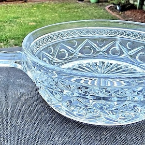 Puede incluir: Cuenco de vidrio transparente con asa. El cuenco presenta un patrón detallado en relieve alrededor del borde y en los lados. El asa es lisa y redondeada. El cuenco está sobre una superficie gris oscuro.