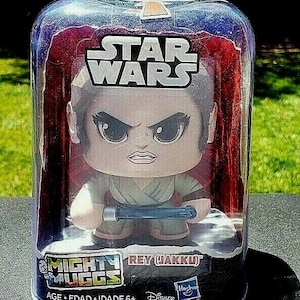 Może przedstawiać: Figurka Mighty Mugg Star Wars Rey (Jakku) w przezroczystej plastikowej obudowie. Figurka ma styl kreskówkowy i trzyma niebieski miecz świetlny. Opakowanie zawiera logo Star Wars i markę Disney.