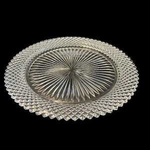 Puede incluir: Un plato de vidrio transparente con un patrón de diamante alrededor del borde y un patrón de sol en el centro.