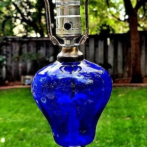 May include: A vintage table lamp with a cobalt blue glass body and a white base. The lamp has a brass-colored metal accent and a green glass finial. The lamp is set against a blurred green background.